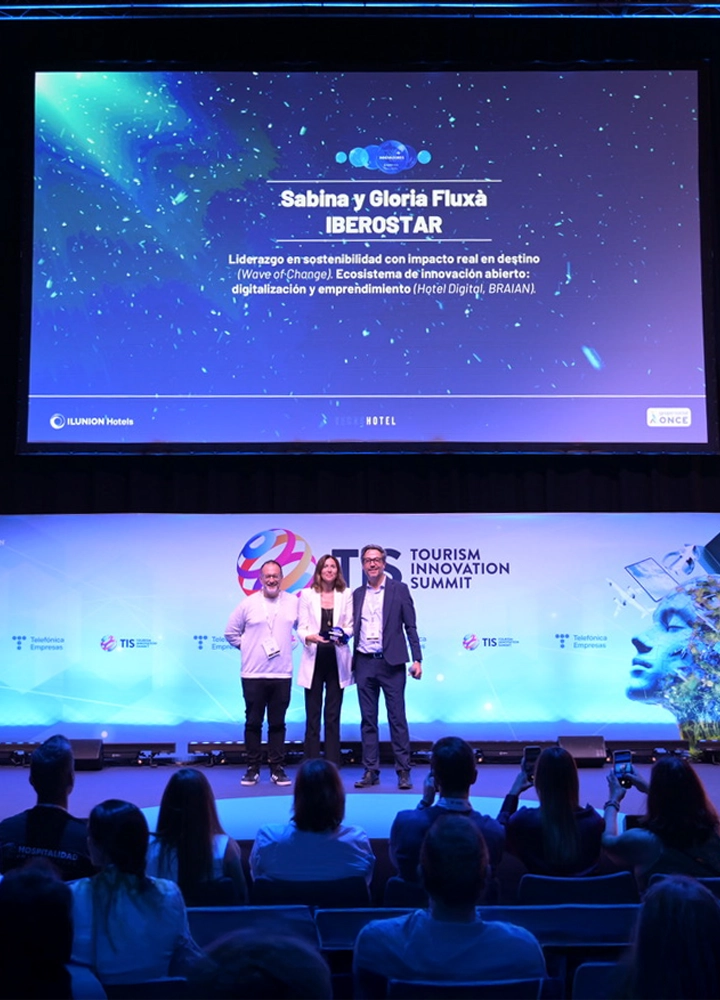 Dos hombres y una mujer de pie en el escenario del Tourism Innovation Summit; la mujer sostiene un premio frente a una pantalla con el nombre Sabina y Gloria Fluxà de Iberostar.