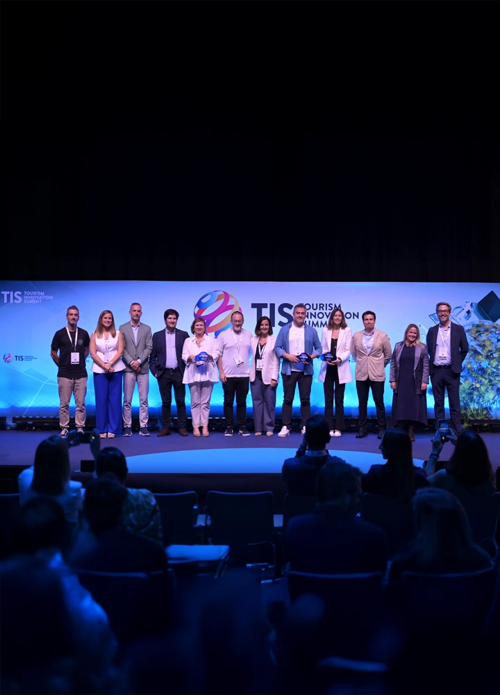 Grupo de personas en el escenario del Tourism Innovation Summit, posando frente a un fondo azul con el logo del evento y público en primer plano.