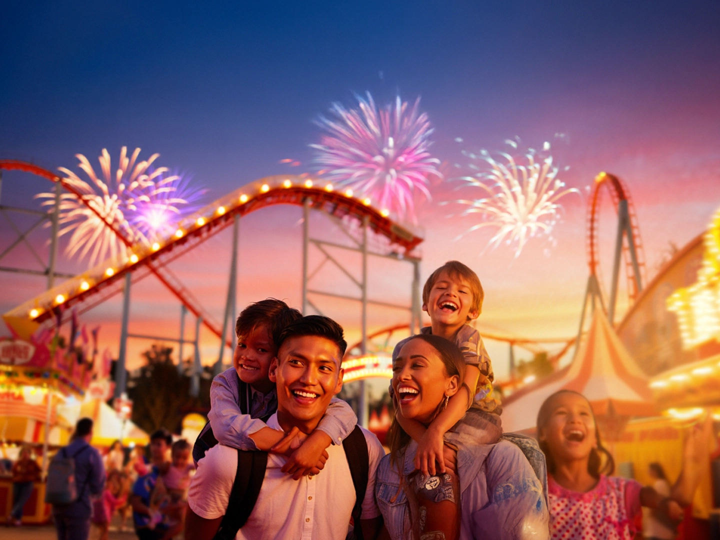 Grupo de personas disfrutando en parque de atracciones al atardecer, con montaña rusa iluminada y fuegos artificiales en el cielo