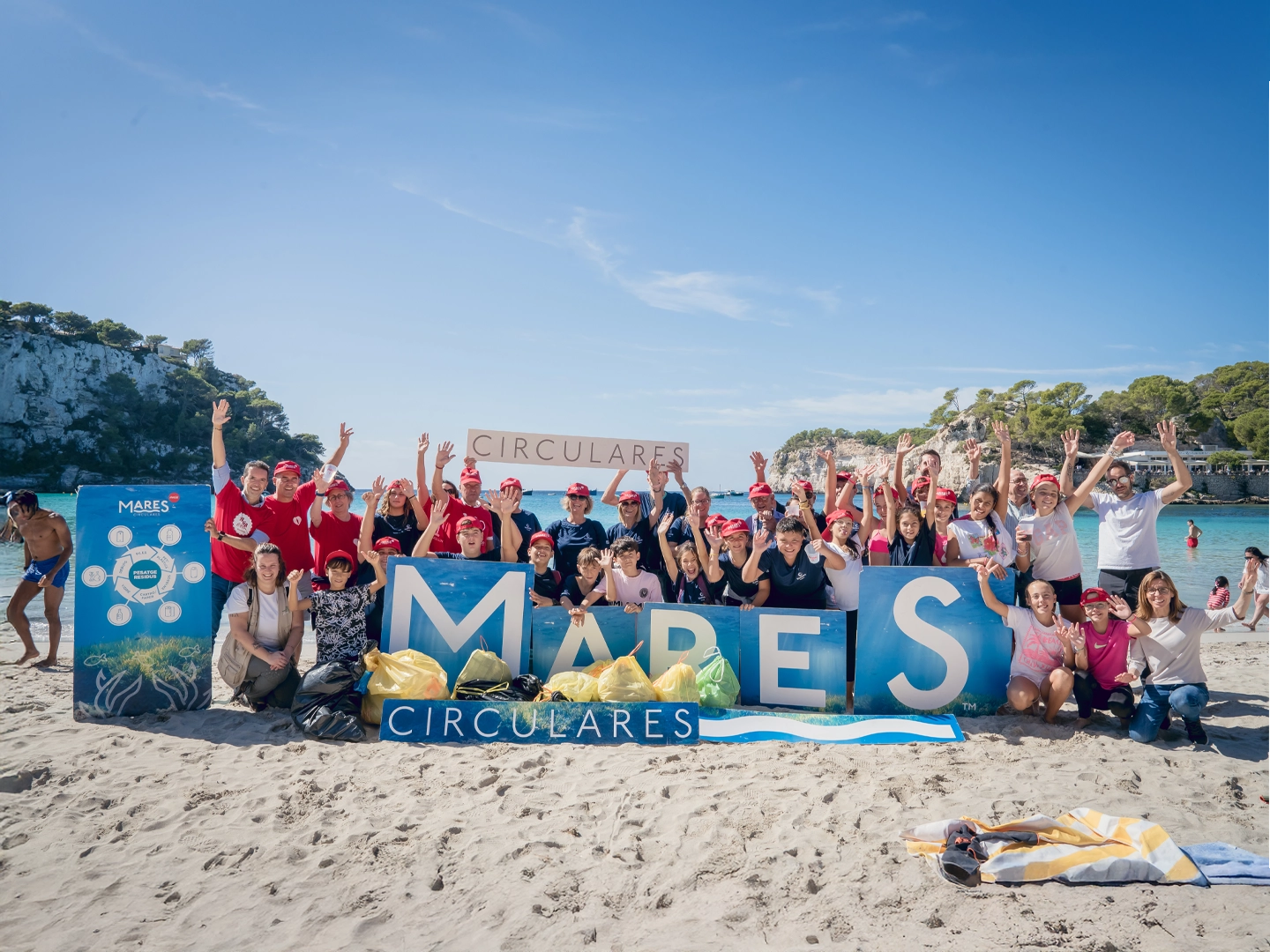 Grupo en playa con bolsas recolectadas, sostienen letras 'MARES' y pancarta 'CIRCULARES' en jornada de limpieza ambiental.