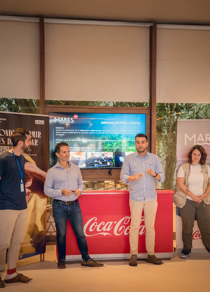 Four people in front of Coca-Cola counter and screen showing 'MARES' and event info, during a presentation with banners on both sides.