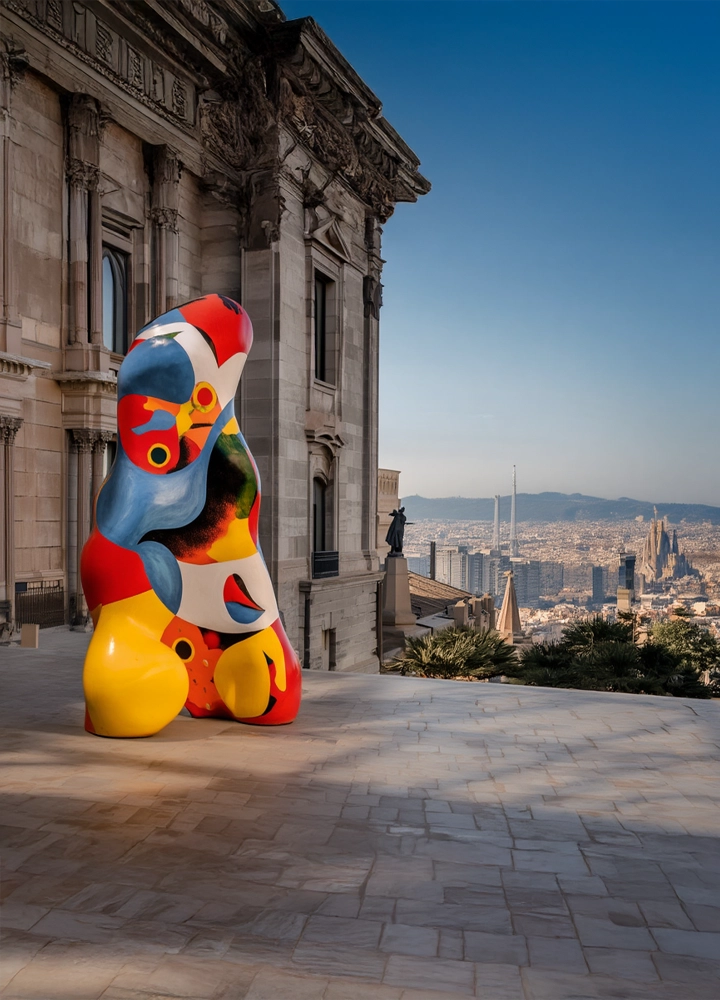 Una escultura abstracta y colorida, con formas orgánicas en rojo, azul, amarillo y negro, se encuentra en una terraza de piedra frente a un edificio clásico y un horizonte urbano.