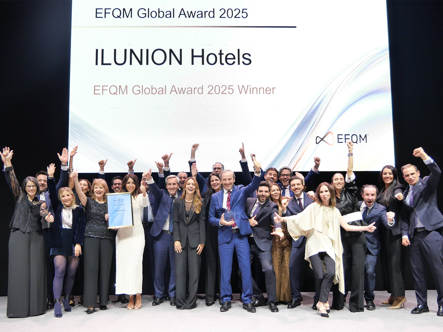 Grupo de personas celebrando en el escenario con un diploma frente a una pantalla que anuncia EFQM Global Award 2025 para ILUNION Hotels.