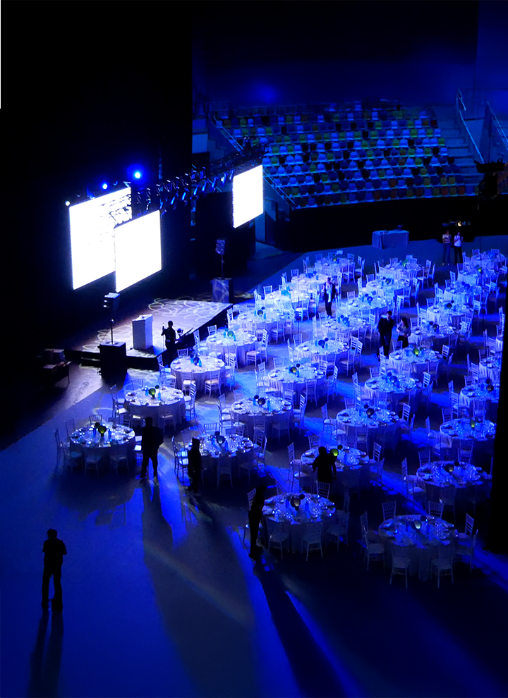Vista aérea de un salón iluminado en azul con mesas redondas preparadas para una gala y pantallas grandes al fondo.