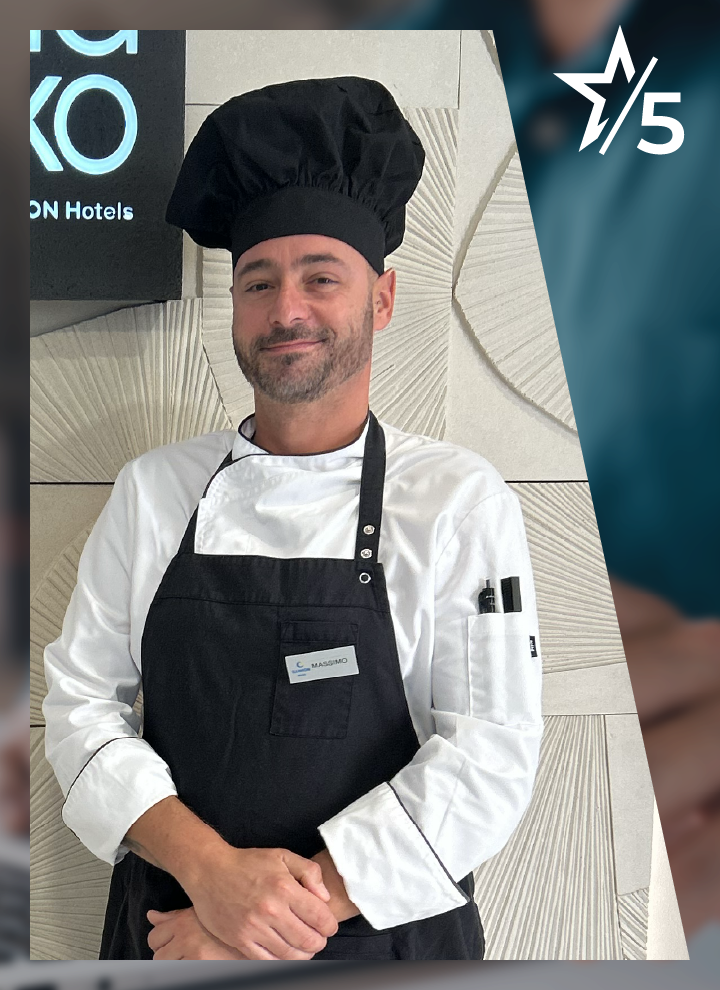 Un hombre con uniforme de cocina y gorro negro, llevando delantal y placa identificativa, posando frente a pared decorativa junto a cartel de ILUNION Hotels.