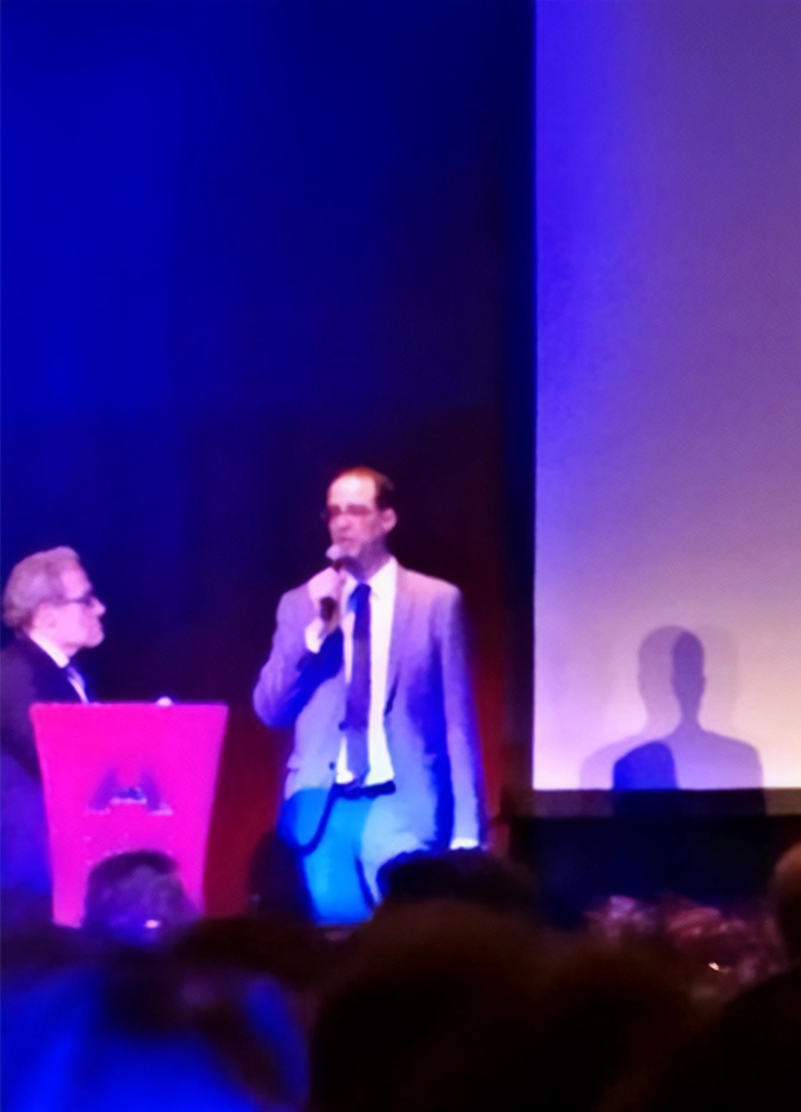 Two people on stage: one speaking into a microphone, the other behind a red podium. Blue background with shadows, presentation setting.