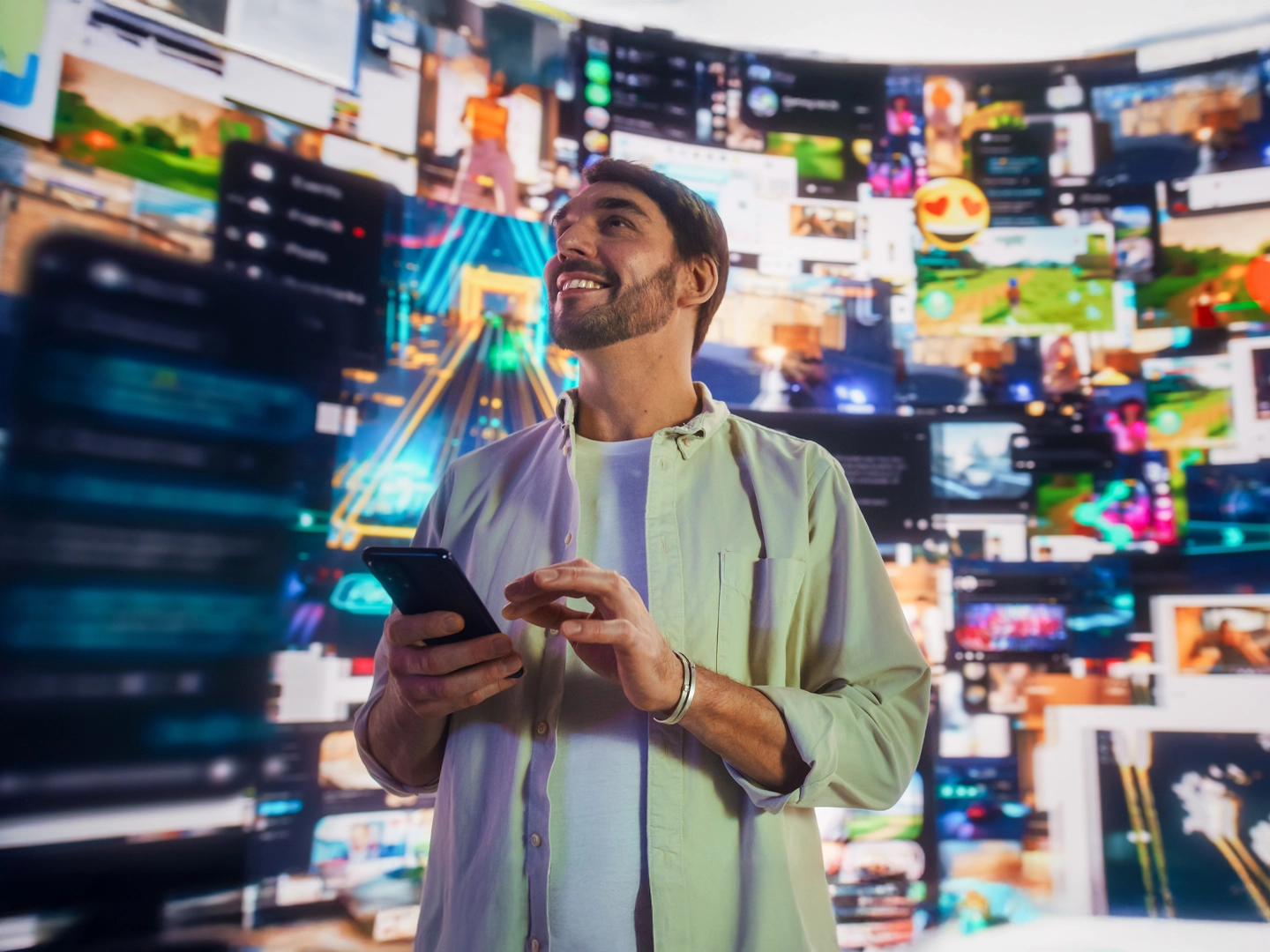 Un hombre sonriente de barba y camisa clara, mira hacia arriba mientras usa un smartphone. Detrás, una pared curva con múltiples pantallas mostrando diversos contenidos digitales.