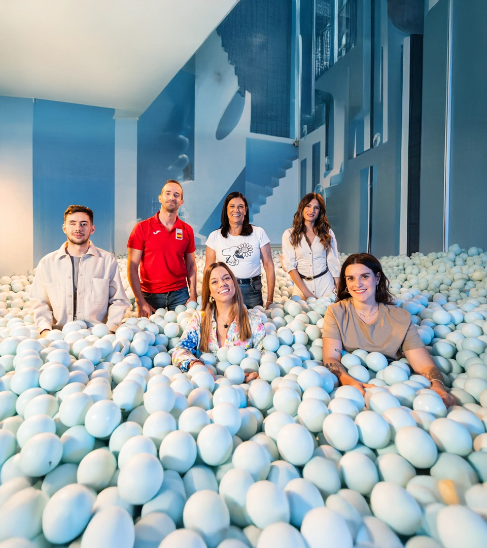 Seis personas dentro de una piscina de bolas, rodeadas por un fondo azul y una estructura moderna que enmarca la escena.