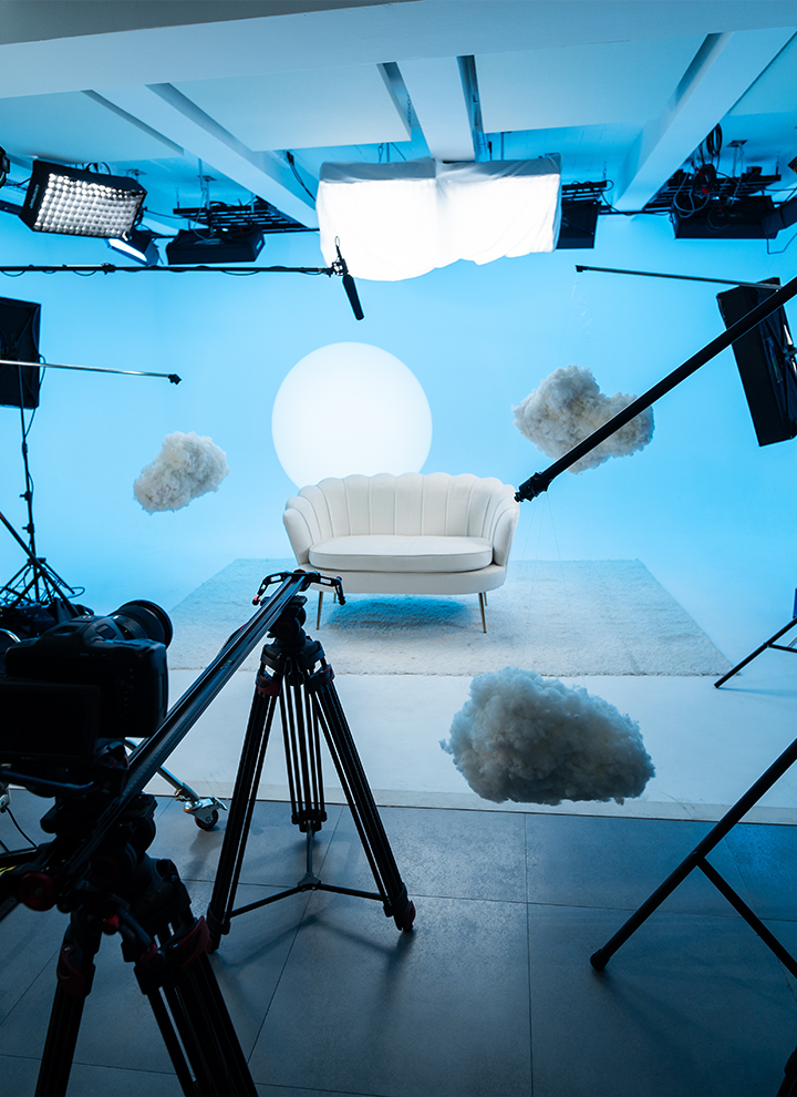 Set de estudio con sofá blanco, nubes suspendidas y fondo azul cielo, iluminado por luz circular y rodeado de equipos de grabación.