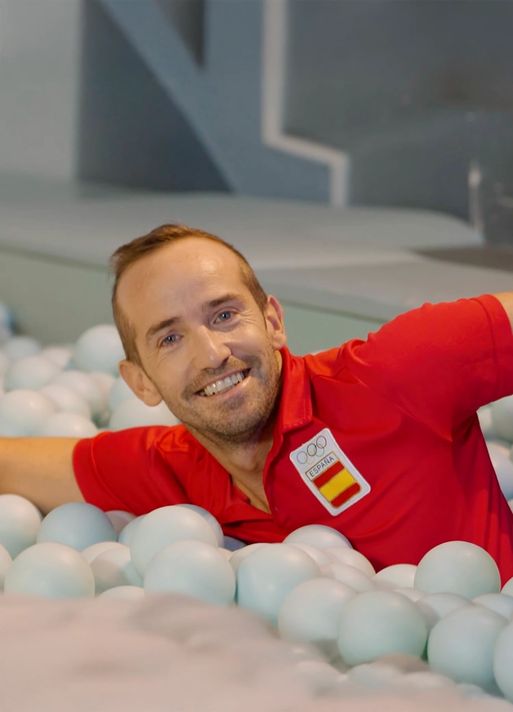 Hombre con polo rojo con insignia olímpica de España sonríe mientras posa en una piscina de bolas celestes.