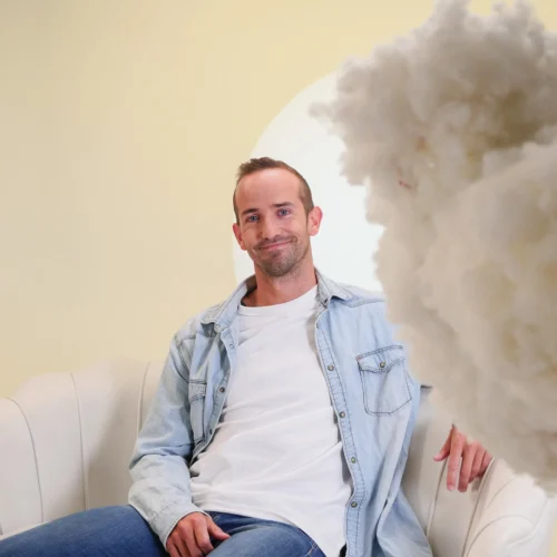 Hombre sentado en sillón blanco con camisa vaquera y camiseta blanca, en fondo claro con objeto esponjoso desenfocado en primer plano.