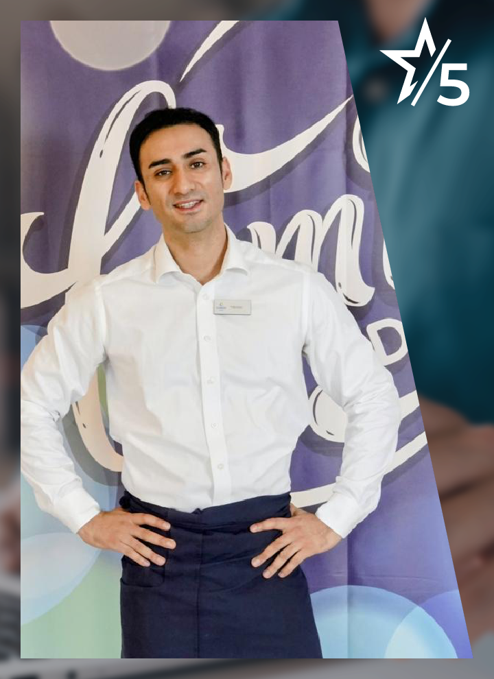 Hombre vestido con camisa blanca y delantal oscuro frente a un fondo decorativo con texto parcial y un símbolo de estrella con número 5.