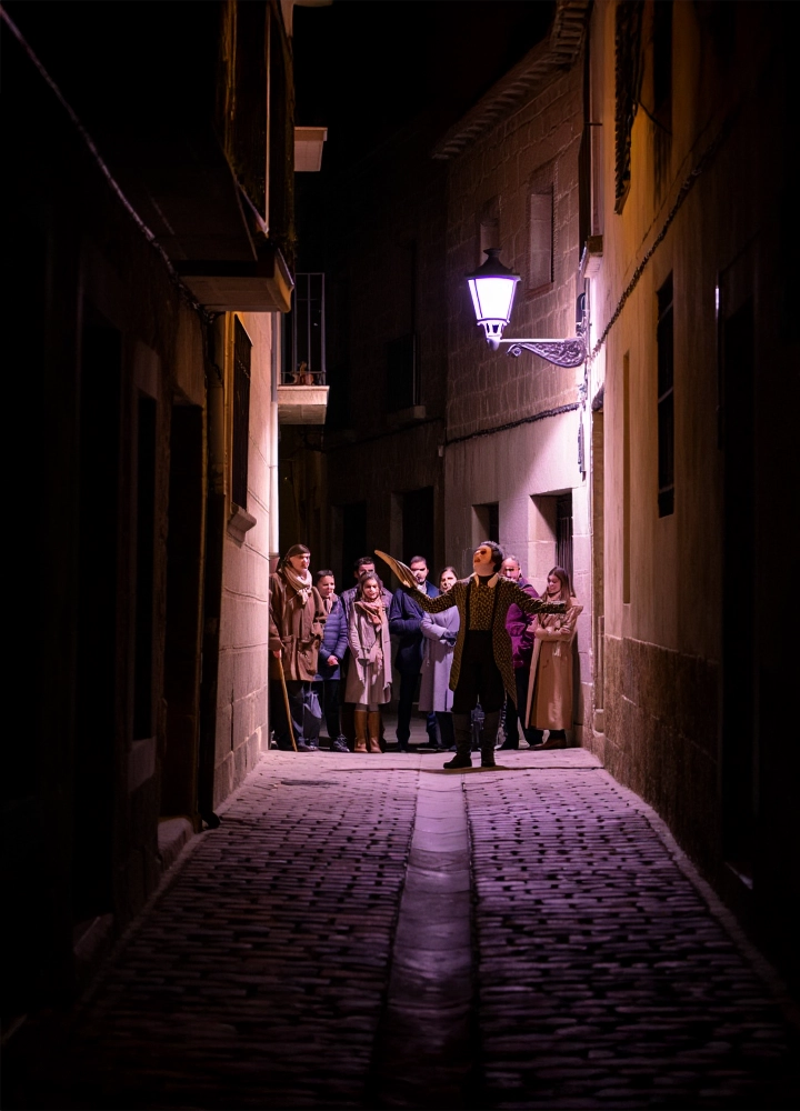 Paseo nocturno por callejón oscuro y empedrado. Un guía con vestimenta de época dirige a un grupo de turistas, iluminado por una farola en la pared.