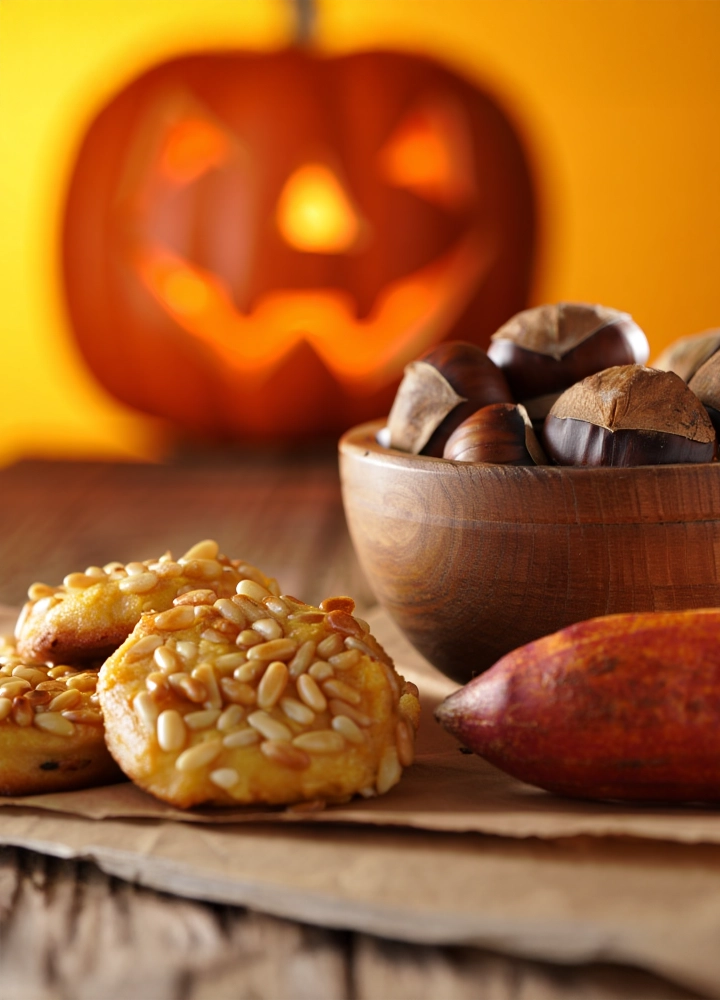 Mesa con dulces de piñones y una batata. Un cuenco de madera contiene castañas. Al fondo, borrosa, brilla una calabaza tallada de Halloween.