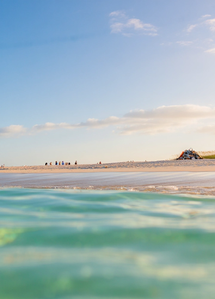 Primer plano del mar turquesa en la mitad inferior de la imagen. Por encima de la línea de agua, una playa de arena blanca con dunas y un cielo azul con nubes blancas.