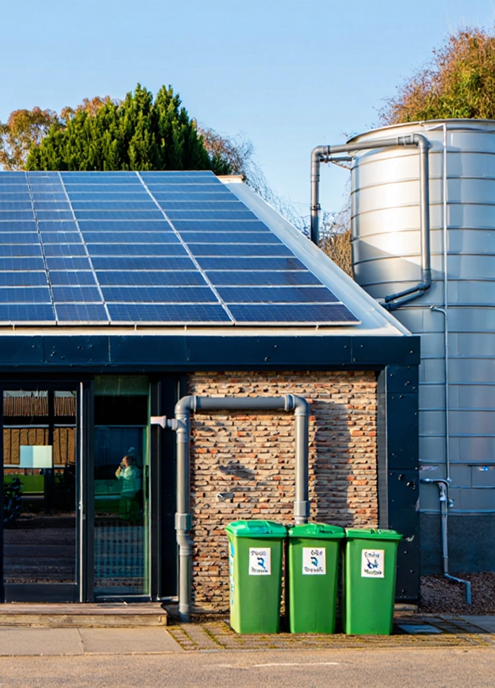 Un edificio con paneles solares en el tejado y un gran depósito de agua gris adyacente. Tres cubos de basura verdes se alinean frente a una pared de ladrillo y una ventana.