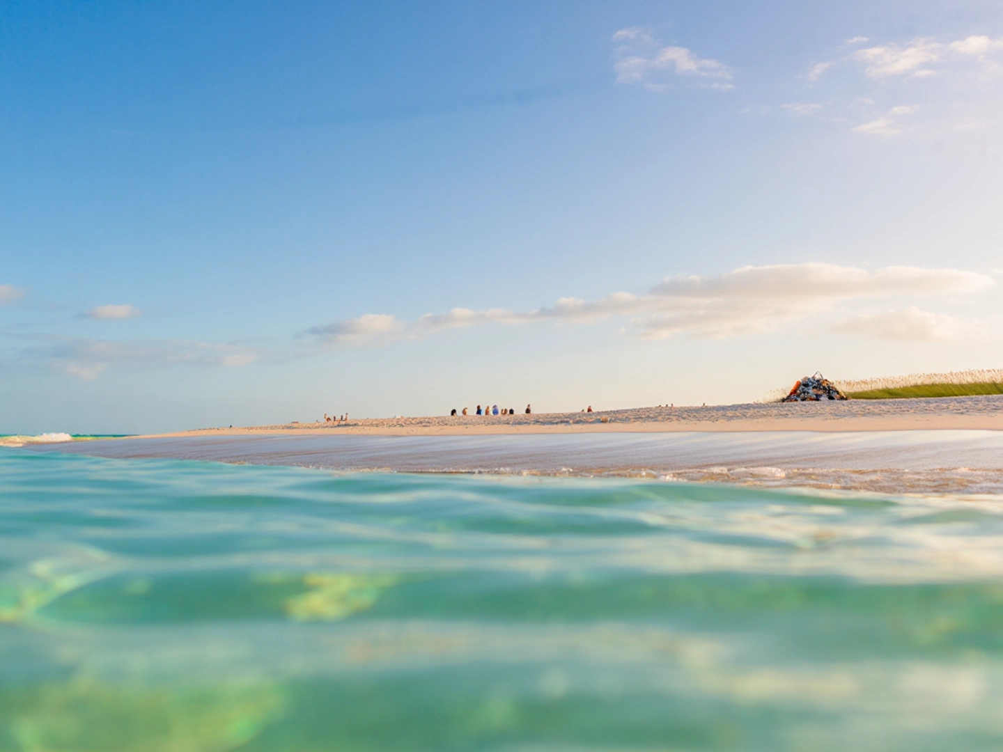 Primer plano del mar turquesa en la mitad inferior de la imagen. Por encima de la línea de agua, una playa de arena blanca con dunas y un cielo azul con nubes blancas.