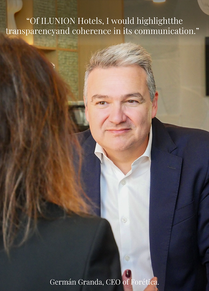 A women and a man seated indoors at a table, the woman facing the camera and the man with back turned, in a professional setting. Upper text reads: ‘Of ILUNION Hotels, I would highlight the transparency and coherence in its communication.’ Lower text reads: ‘Germán Granda, CEO of Forética.’