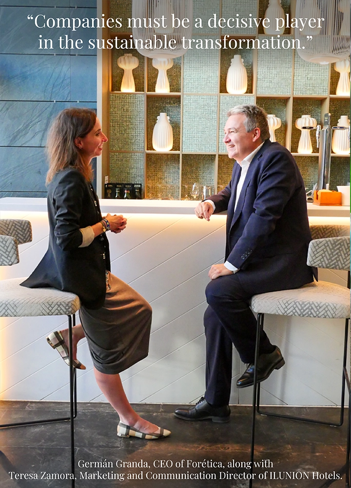 A man and a woman seated on bar stools in a modern setting with a decorative shelf and warm lighting. Upper text reads: ‘Companies must be a decisive player in the sustainable transformation.’ Lower text reads: ‘Germán Granda, CEO of Forética, along with Teresa Zamora, Marketing and Communication Director of ILUNION Hotels.’