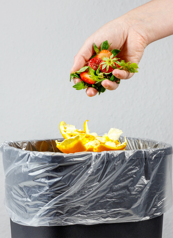 Mano sosteniendo restos de fresa sobre cubo de basura con cáscaras de naranja, ilustrando el desecho de residuos orgánicos.