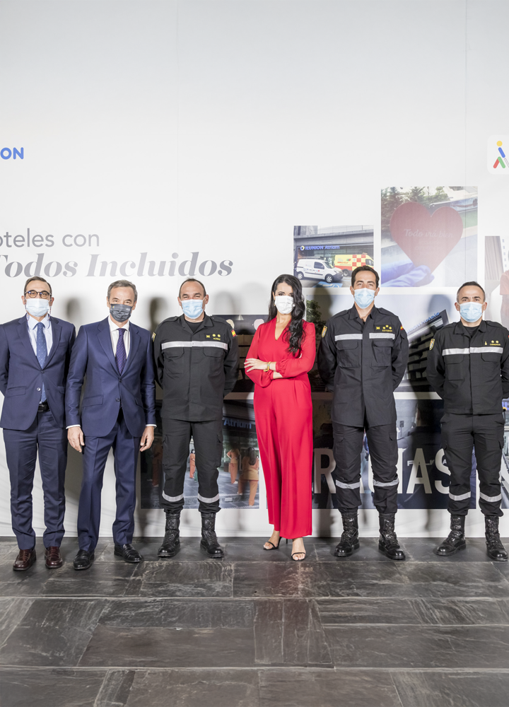 Six people standing in a row; three in dark uniforms and three in formal attire, one in red. Background with the text 'Hotels with Everyone Included' and images.