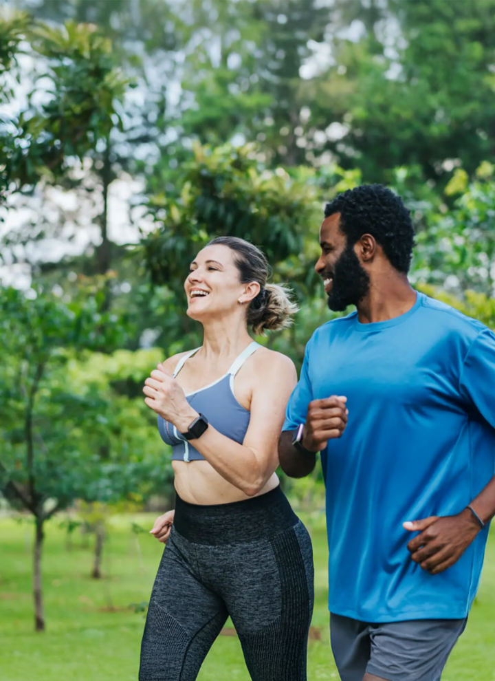 Una mujer y un hombre corren al aire libre en un parque con árboles y césped, vestidas con ropa deportiva, en un entorno natural y saludable.