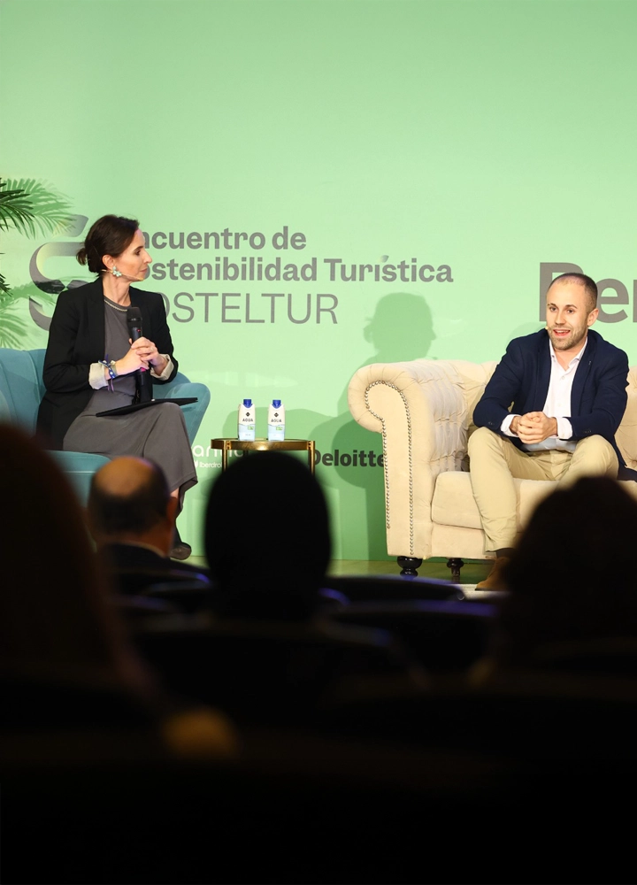 Un hombre y una mujer sentadas en sillones sobre un escenario con fondo verde que muestra el texto Encuentro de Sostenibilidad Turística HOSTELTUR, junto a botellas de agua y logotipo de Deloitte.