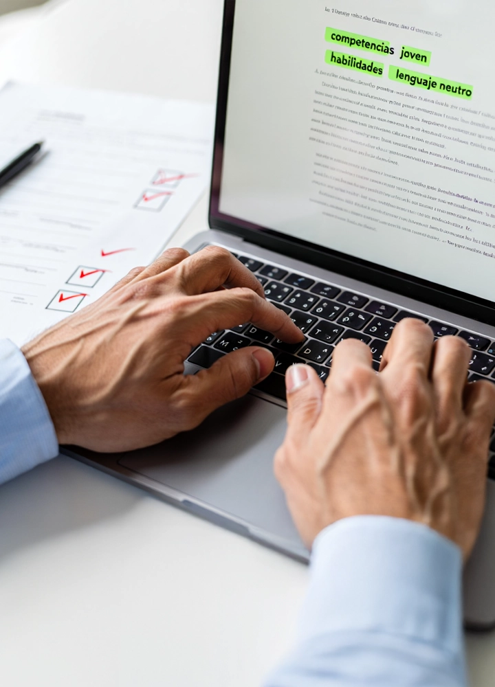 Manos escribiendo en un portátil sobre una mesa, con documento impreso al lado y texto en pantalla que resalta palabras como competencias, habilidades y lenguaje neutro.
