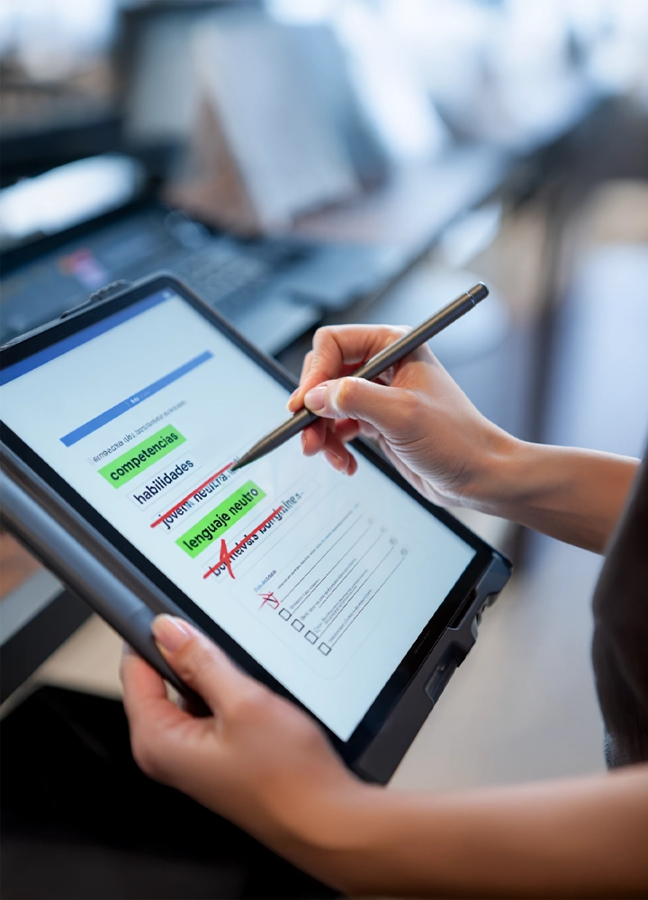 Manos sosteniendo una tableta con un cuestionario en pantalla, usando un lápiz digital para marcar opciones relacionadas con competencias, habilidades y lenguaje neutro.