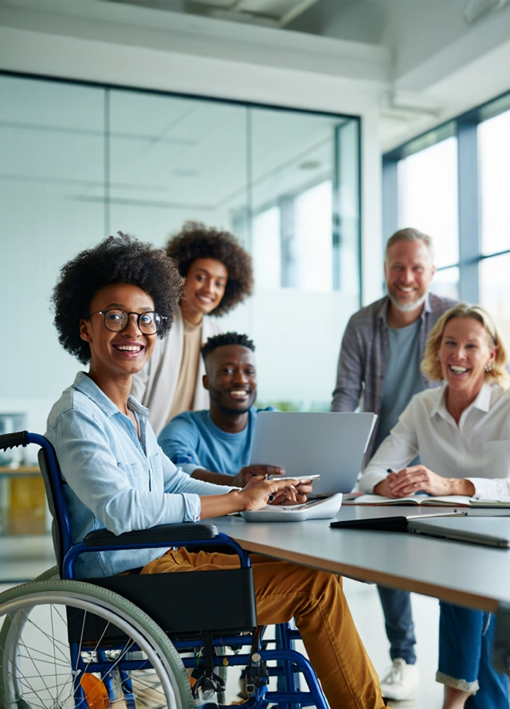 Grupo de cinco personas reunidas en una oficina moderna, sentadas y de pie alrededor de una mesa con portátiles y documentos, incluyendo una persona en silla de ruedas.