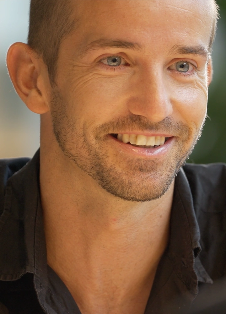 Primer plano de hombre sonriente, vestido con camisa oscura sentado en interior, imagen centrada en la parte superior del torso y cuello.