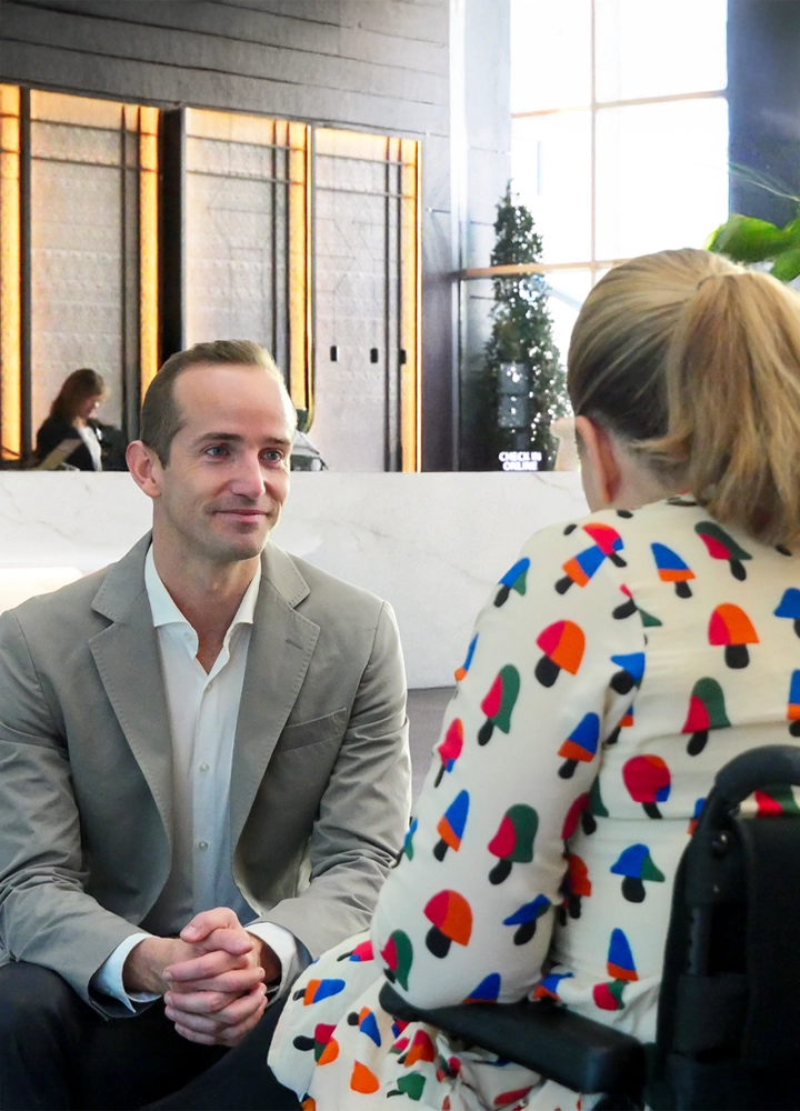 Hombre con traje gris y camisa blanca sentado frente a mujer con vestido estampado en un vestíbulo elegante con mostrador de recepción y decoración moderna.