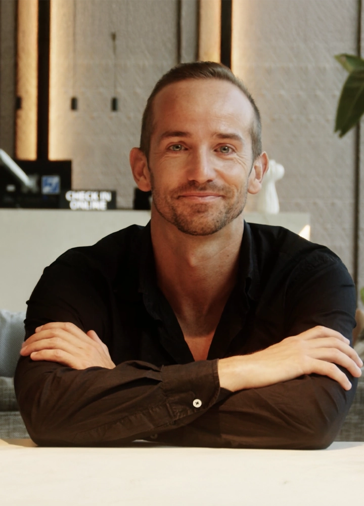 Hombre con camisa negra sentado con brazos cruzados sobre una mesa en un interior moderno, con fondo que incluye columnas y un cartel que dice “Check in Online”.