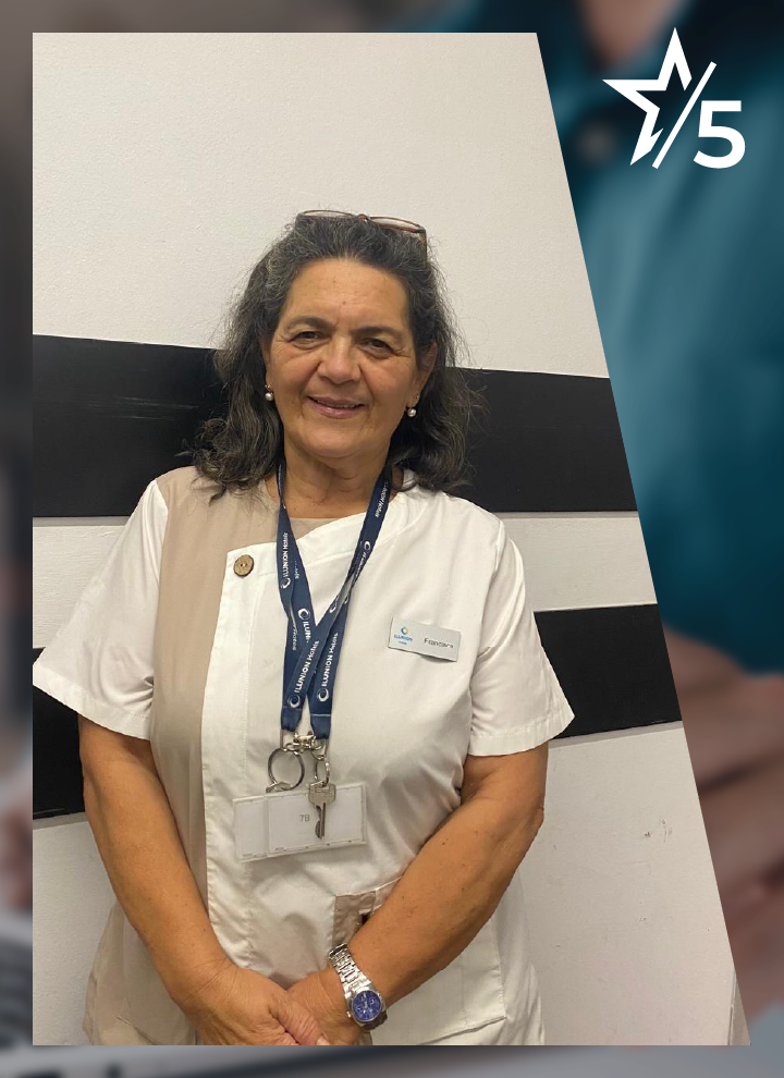 Mujer con uniforme blanco y beige, acreditaciones colgadas al cuello y sonrisa amable, posa frente a pared de franjas negras y blancas.