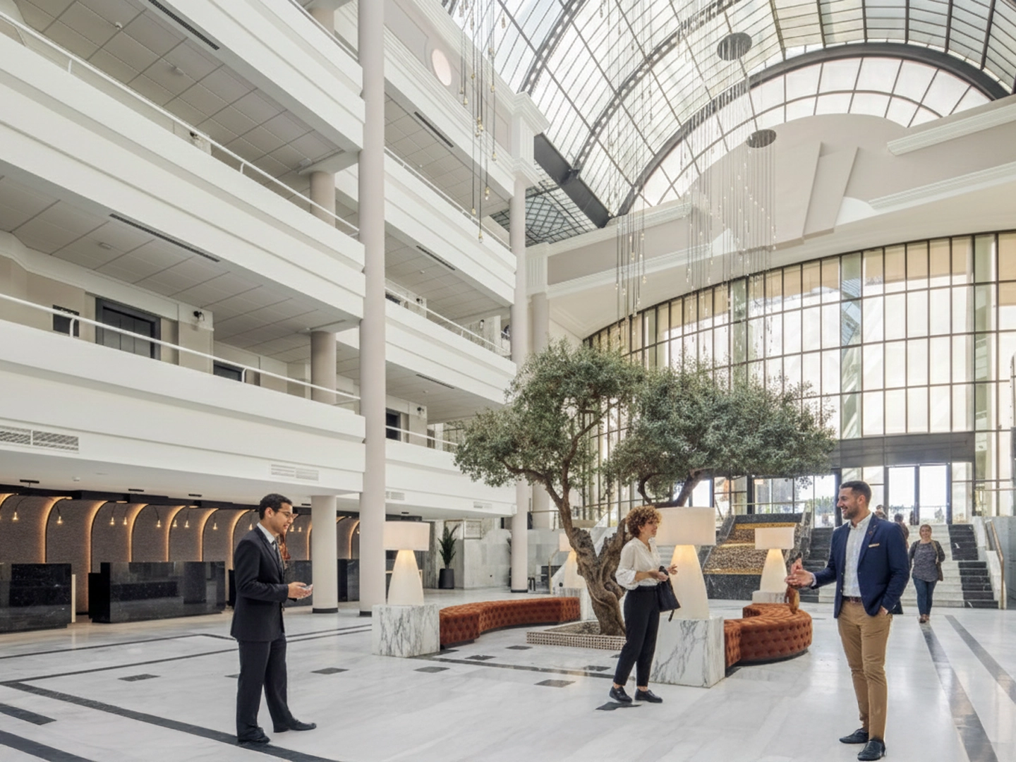 Amplio vestíbulo moderno con techo acristalado, árbol decorativo y lámparas; tres personas conversan y caminan cerca de la zona de recepción del hotel.