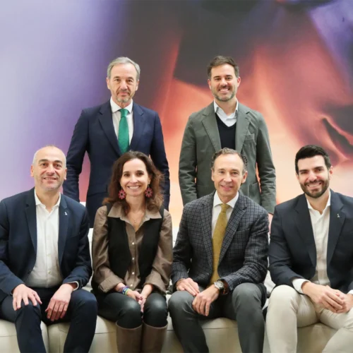 Seis personas posando en grupo sobre un sofá blanco frente a un fondo abstracto con tonos cálidos.