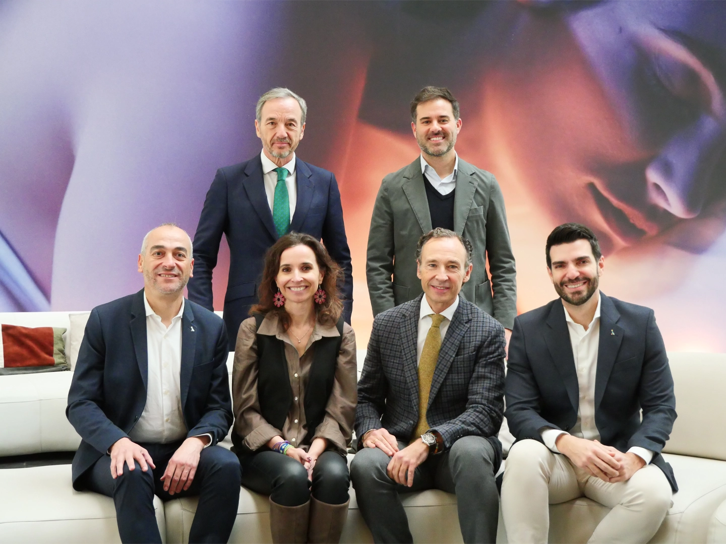 Seis personas posando en grupo sobre un sofá blanco frente a un fondo abstracto con tonos cálidos.