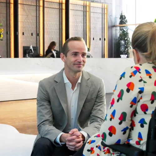 Hombre con traje gris y camisa blanca sentado frente a mujer con vestido estampado en un vestíbulo elegante con mostrador de recepción y decoración moderna.
