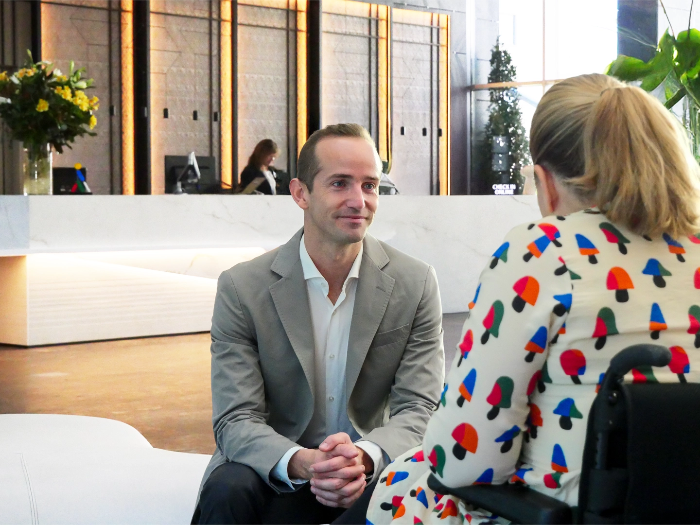Hombre con traje gris y camisa blanca sentado frente a mujer con vestido estampado en un vestíbulo elegante con mostrador de recepción y decoración moderna.