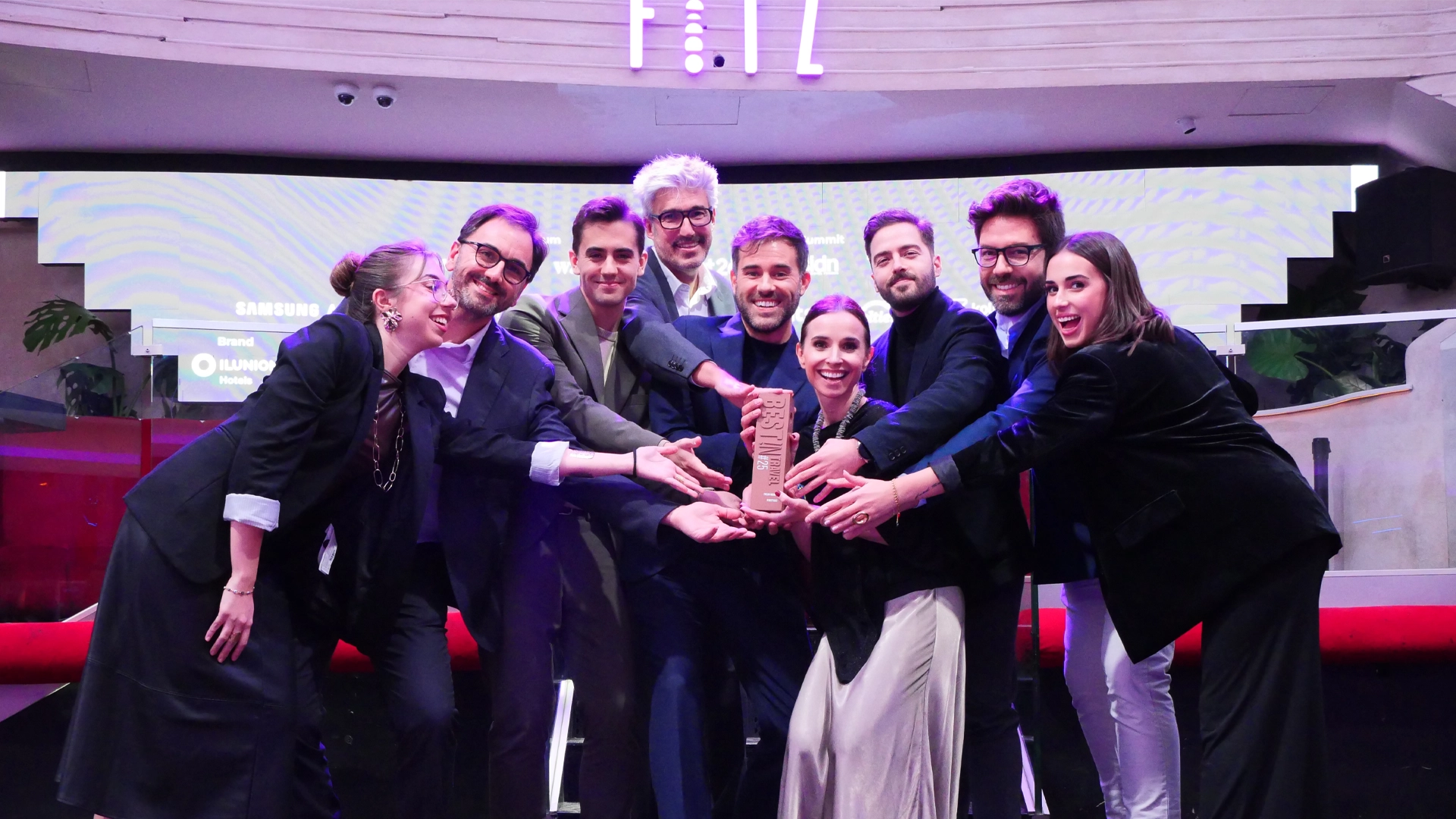 Grupo de ocho personas vestidas de manera formal, reunidas en un escenario interior mientras sostienen un trofeo grande en el centro.