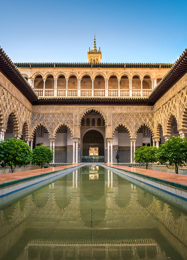 Patio interior del Real Alcázar de Sevilla con arcos decorados, vegetación simétrica y un estanque central reflejando la arquitectura.