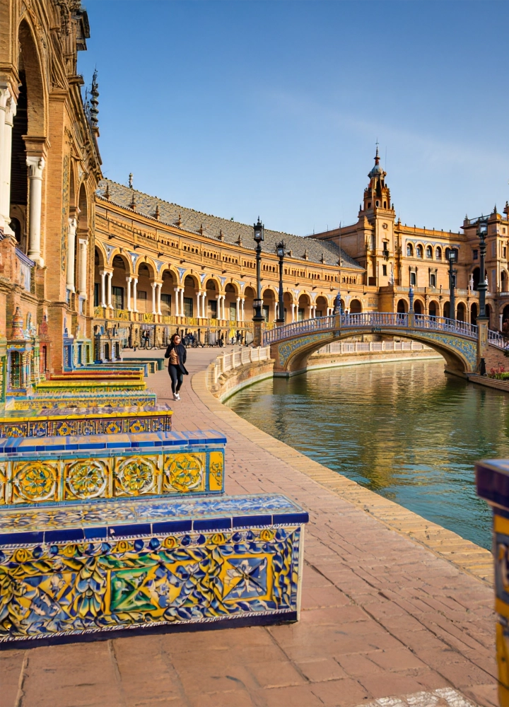 Vista de la Plaza de España en Sevilla con bancos de azulejos coloridos, un canal y una persona caminando junto a los arcos del edificio histórico