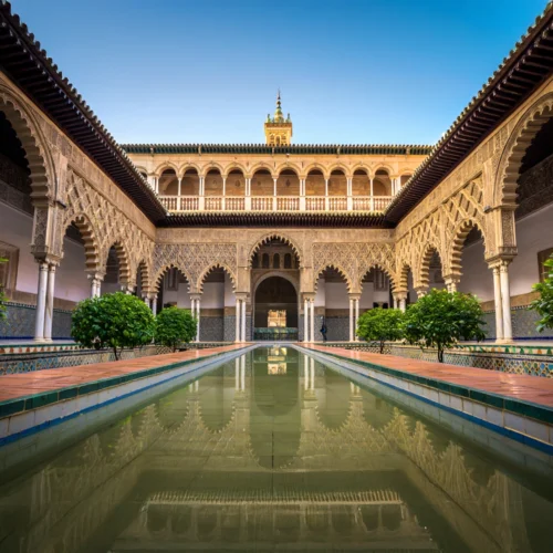 Patio interior del Real Alcázar de Sevilla con arcos decorados, vegetación simétrica y un estanque central reflejando la arquitectura.