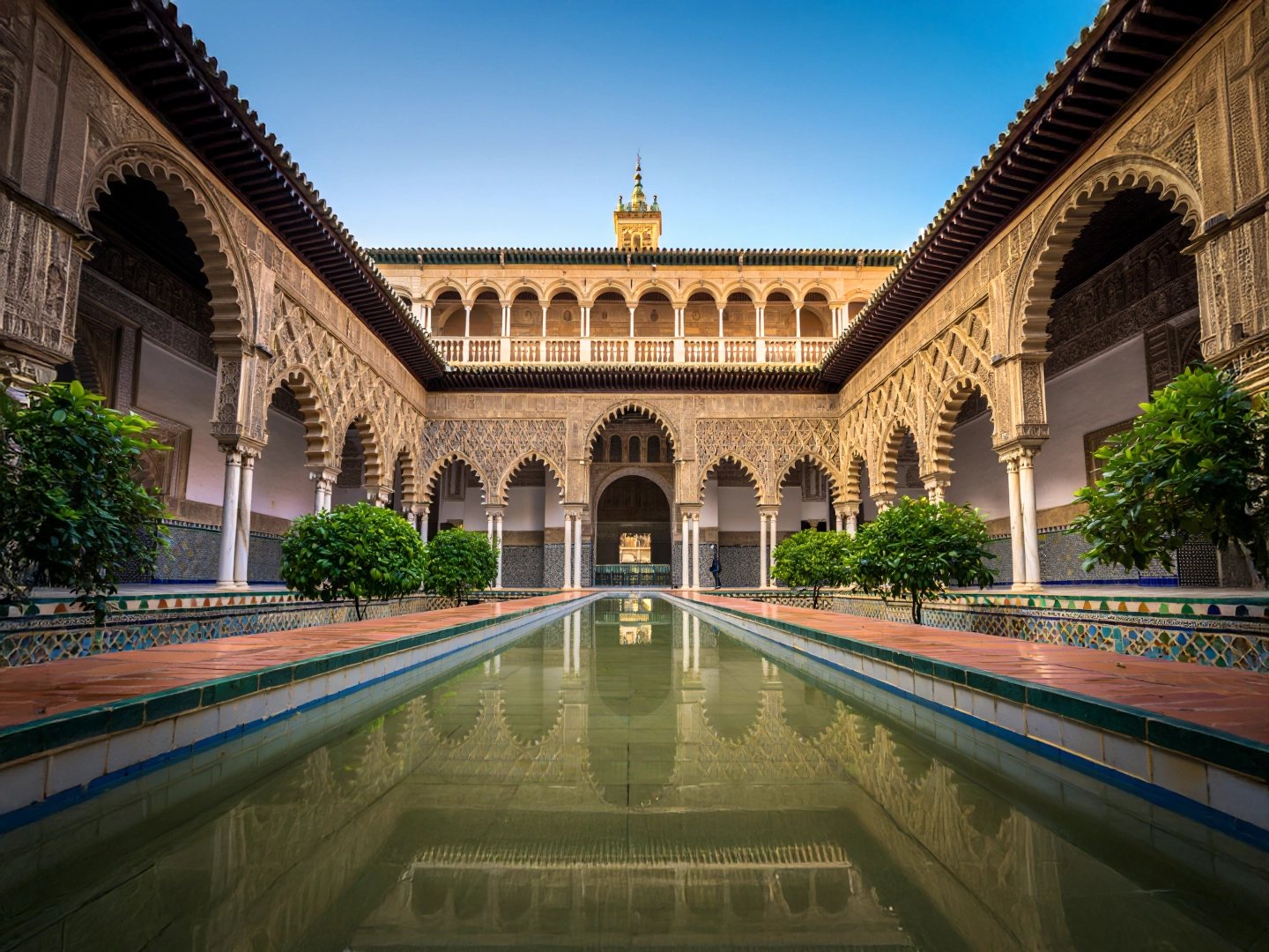 Patio interior del Real Alcázar de Sevilla con arcos decorados, vegetación simétrica y un estanque central reflejando la arquitectura.