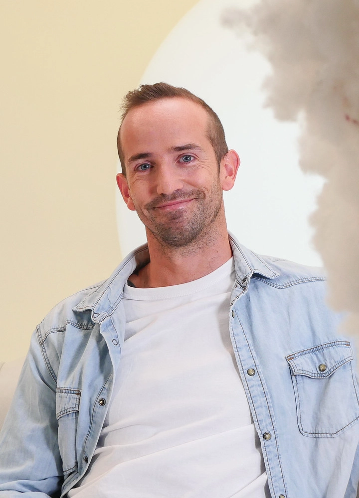 Hombre con camisa vaquera azul claro y camiseta blanca sonríe mientras está sentado en un entorno cálido con una nube decorativa blanca.