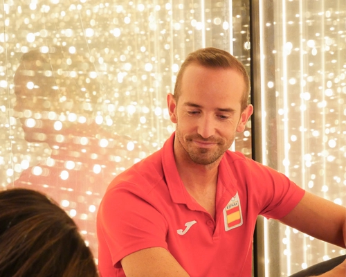 Hombre con polo rojo con la bandera de España posa en un entorno rodeado de luces brillantes.
