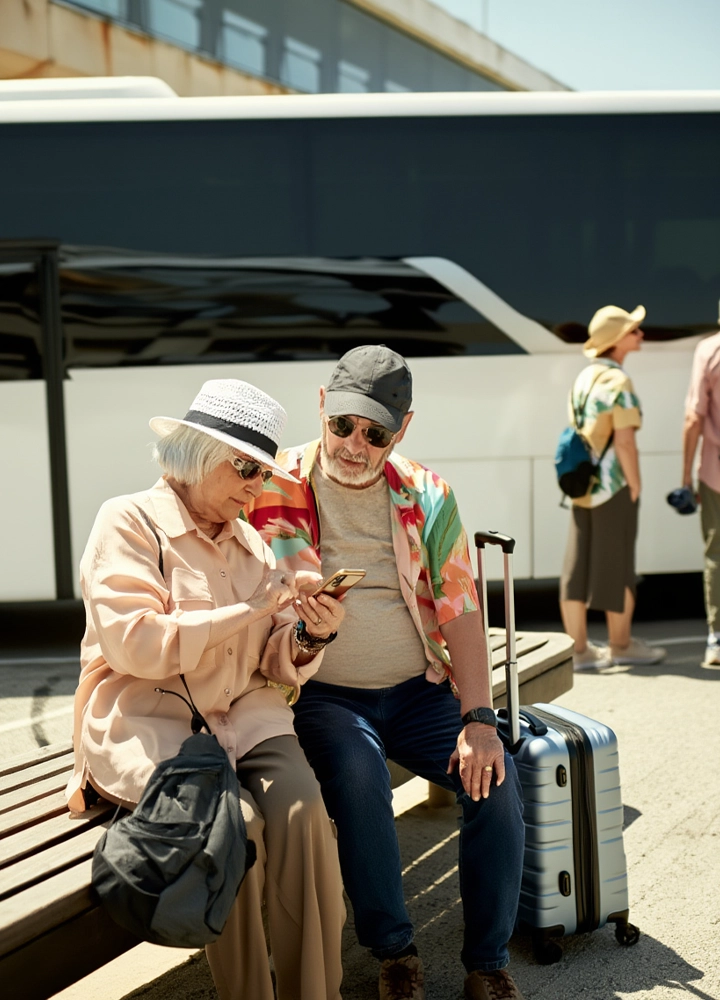 Hombre y mujer sentados en un banco junto a una maleta, mirando un móvil frente a un autobús estacionado en una zona exterior.