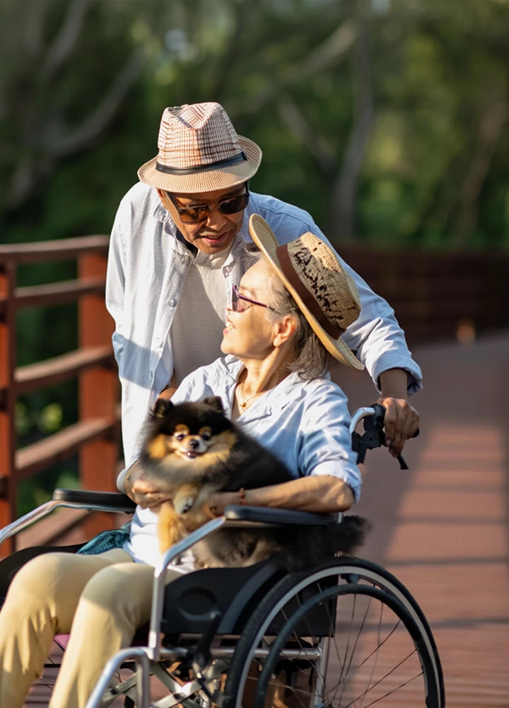 Hombre inclinado junto a una mujer en silla de ruedas que sostiene un perro pequeño, sobre un puente de madera en un entorno natural.