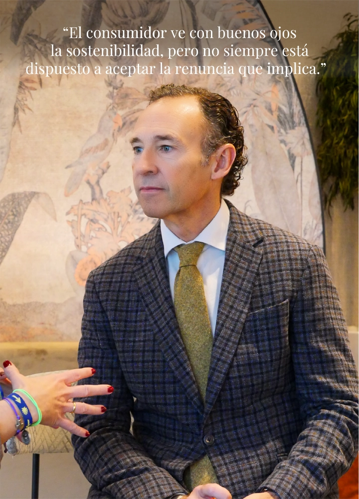 Hombre con chaqueta de cuadros y corbata dorada sentada frente a fondo decorativo; se lee la frase: 'El consumidor ve con buenos ojos la sostenibilidad, pero no siempre está dispuesto a aceptar la renuncia que implica.