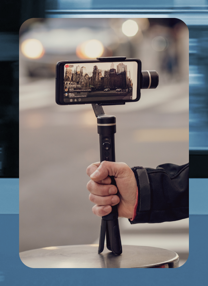 Mano sosteniendo un estabilizador con un móvil grabando una calle urbana con edificios al fondo.